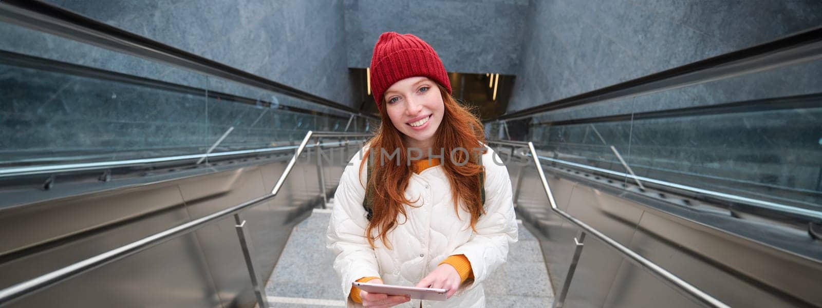 Beautiful redhead female model posing in city, walking up stairs with digital tablet, using gadget to plan her route, reading while going somewere by Benzoix