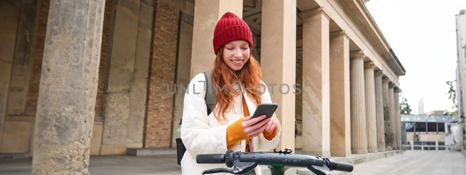 Beautiful young woman, tourist with backpack, rents e-scooter to travel around town, uses mobile phone application.