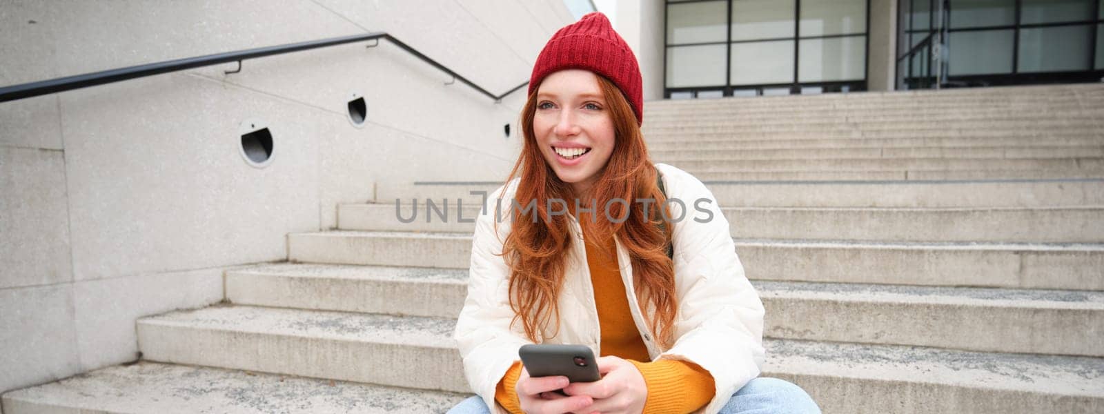 Redhead girl in red hat, sits on stairs and uses mobile phone. Modern woman holding smartphone, texting message, using telephone application outdoors.