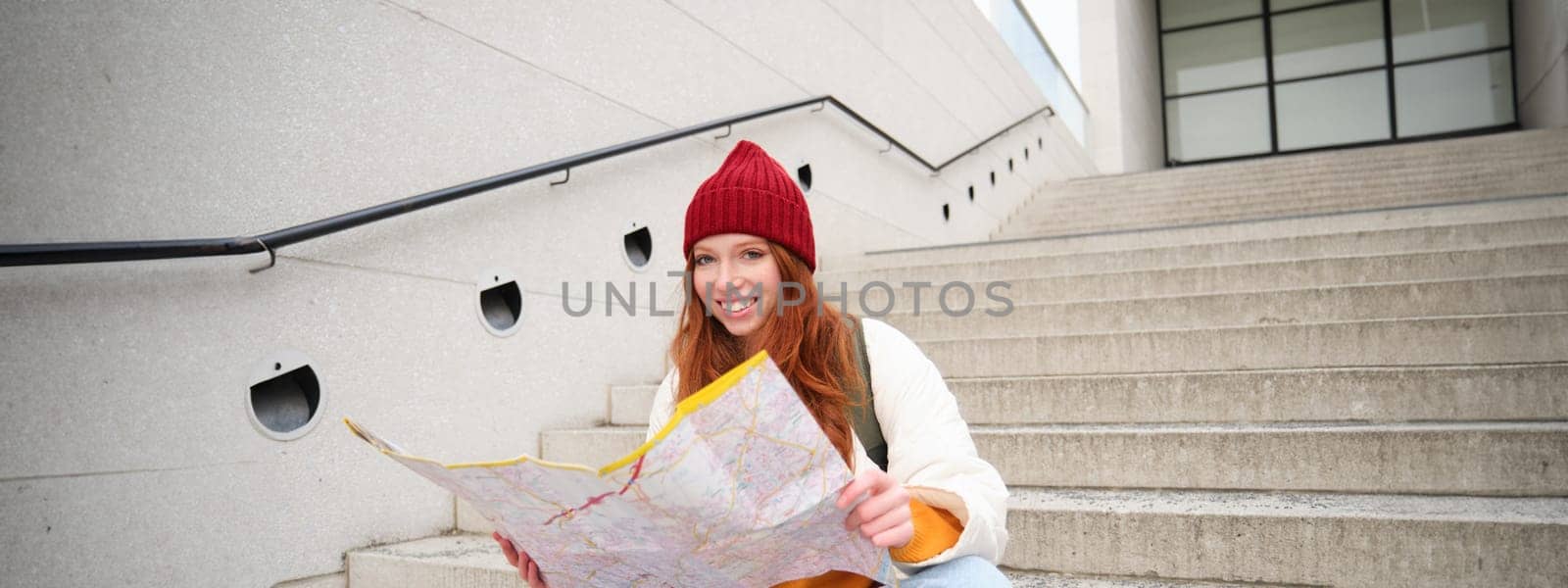 Beautiful girl tourist sits on stairs with city map, plans her journey, looks for direction while travelling around town, searches route for sightseeing.