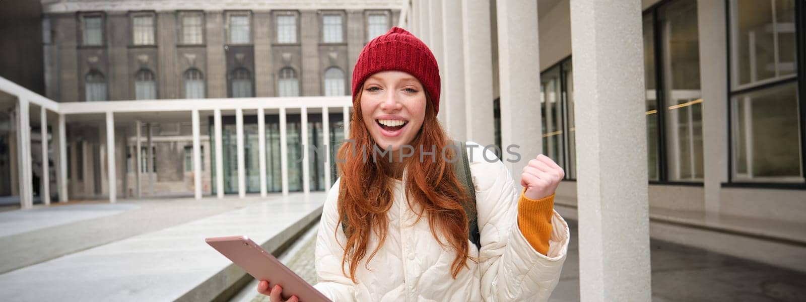 Stylish ginger girl, tourist walks with digital tablet around city, woman connects to iternet on her gadget, looking up information, texting message.