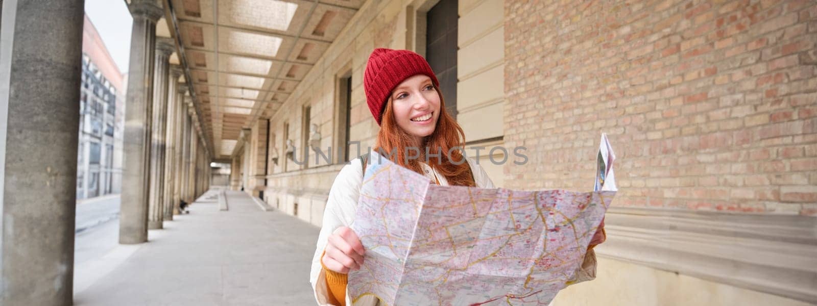 Smiling young redhead woman in red hat, looks at paper map to look for tourist attraction. Tourism and people concept. Girl explores city, tried to find way.