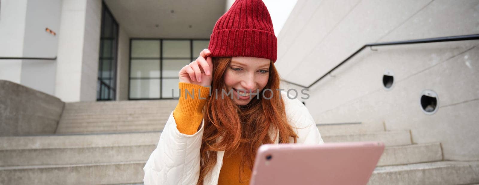 Beautiful redhead female model, ginger girl with digital tablet, sits on stairs outdoors, reads on her gadget, uses internet application and wifi on street by Benzoix