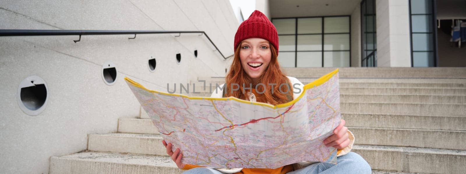 Beautiful girl tourist sits on stairs with city map, plans her journey, looks for direction while travelling around town, searches route for sightseeing.