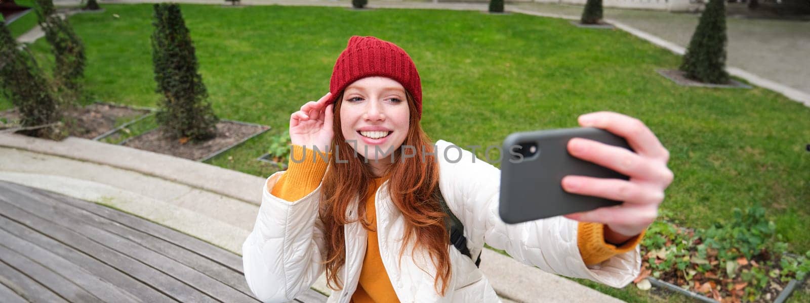 Beautiful young urban girl takes selfie on smartphone, relaxed on bench in park and makes a photo of herself with mobile phone camera.