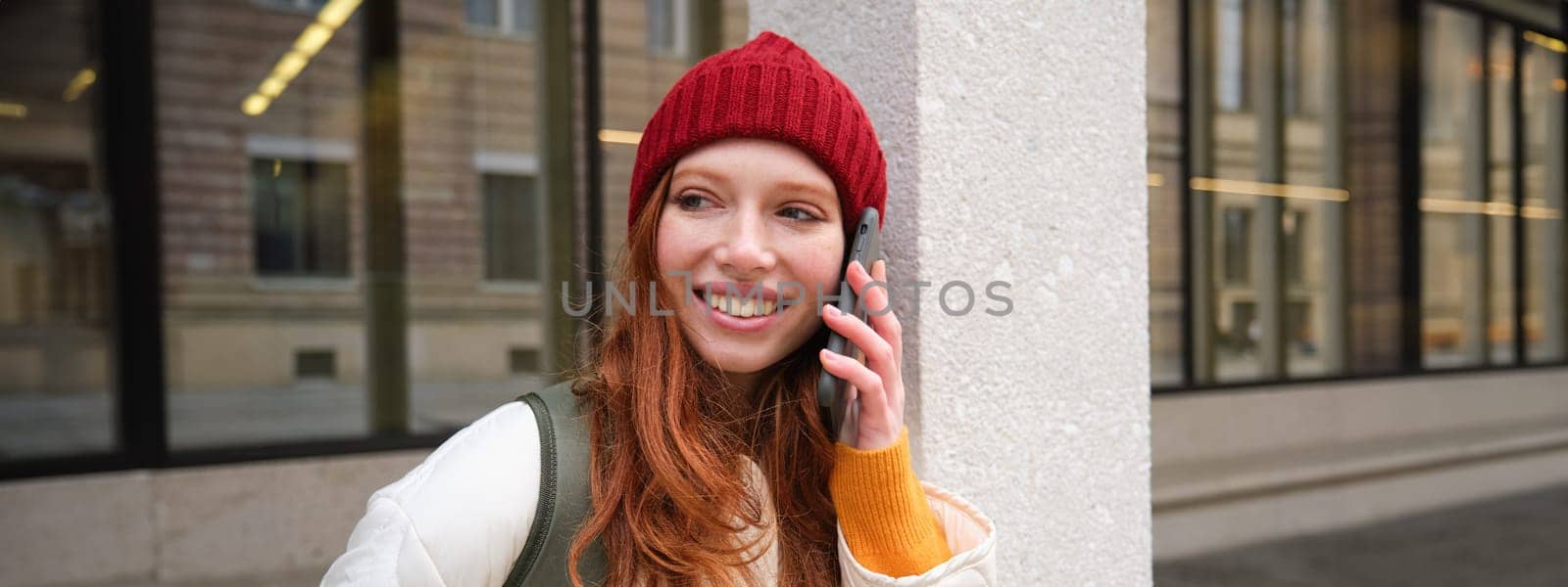 Young people and mobile connection. Happy redhead girl talks on phone, makes telephone call, stands outdoors with backpack and uses smartphone app.