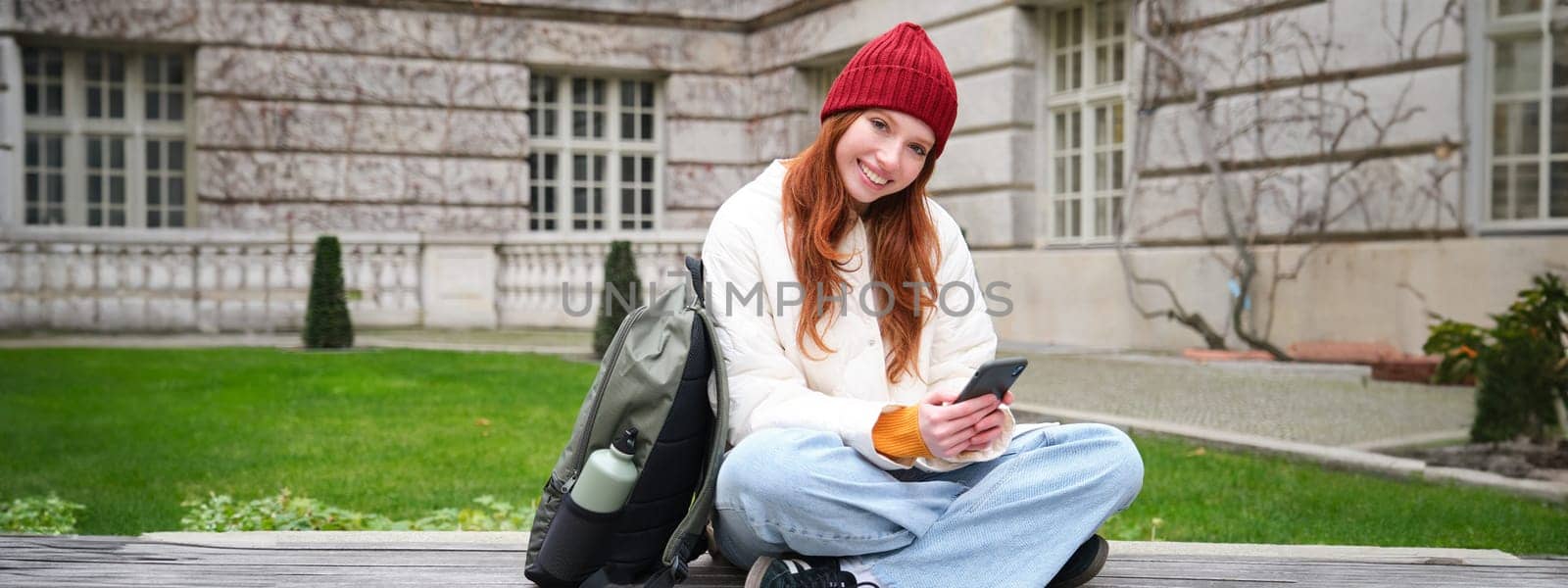 Redhead girl, female student sits with mobile phone on bench in parj, leans on her backpack. Woman browsing social media app feed on her smartphone.