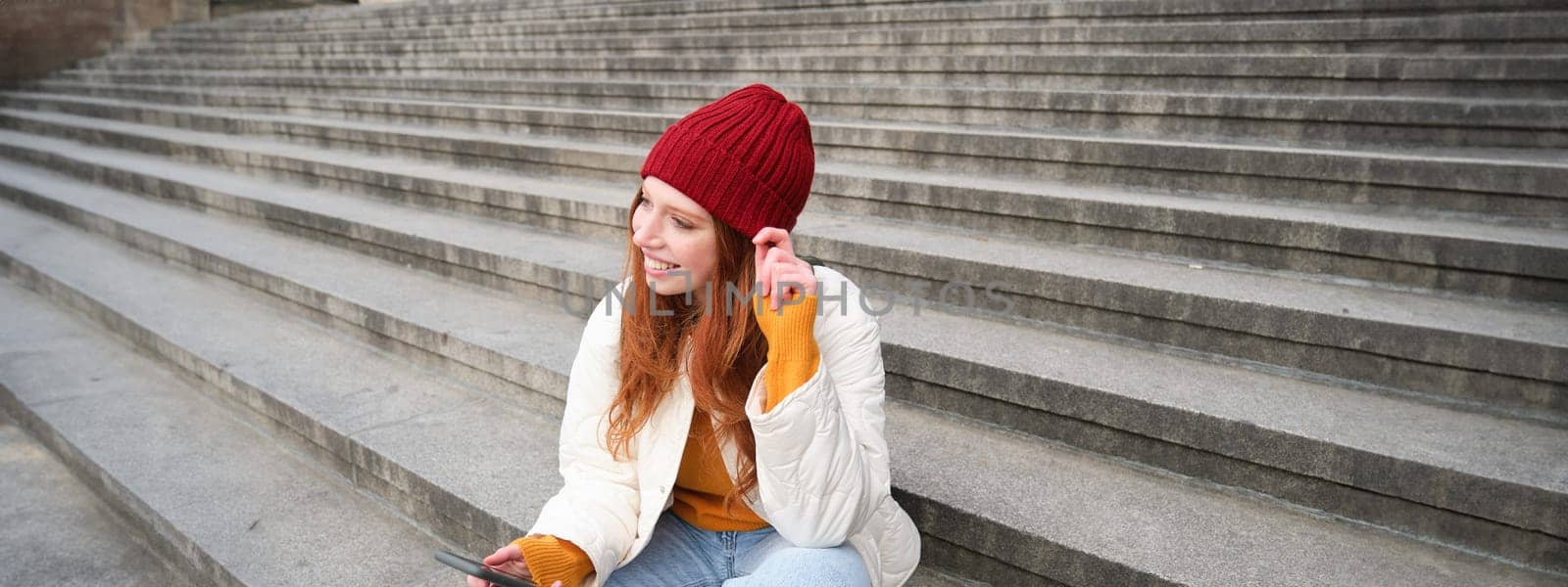 Portrait of young urban girl in red hat, sits on stairs near museum, holds mobile phone, connects to public wifi and surfs net, uses smartphone apps.