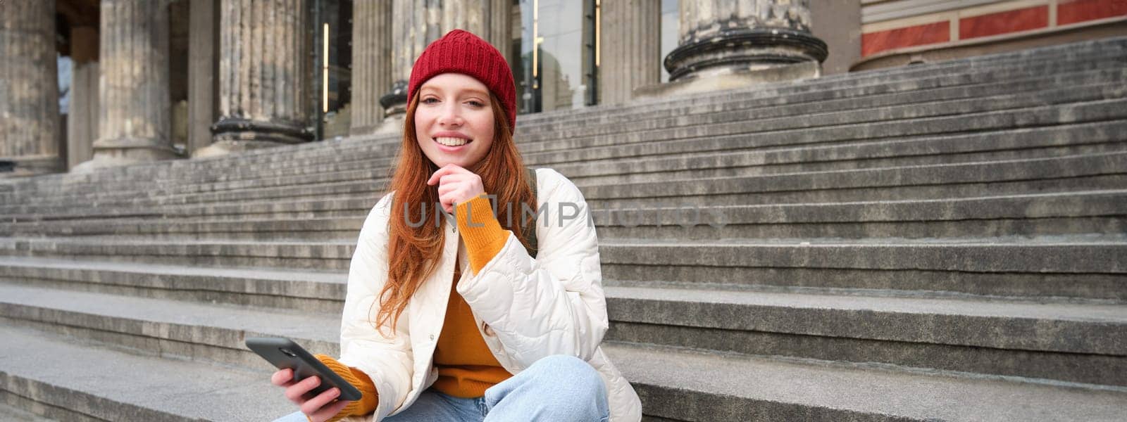 Stylish young redhead woman, talking on mobile phone app, using social media application, looking for something online on smartphone, sits on stairs outdoors.