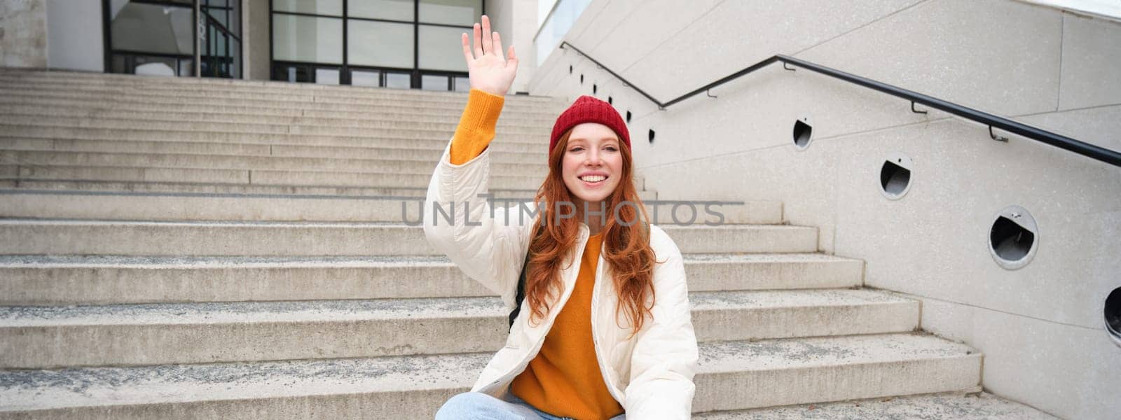 Smiling young redhead girl waits someone on stairs outdoors, waves hand at friend, holds smartphone, says hello. People and lifestyle concept