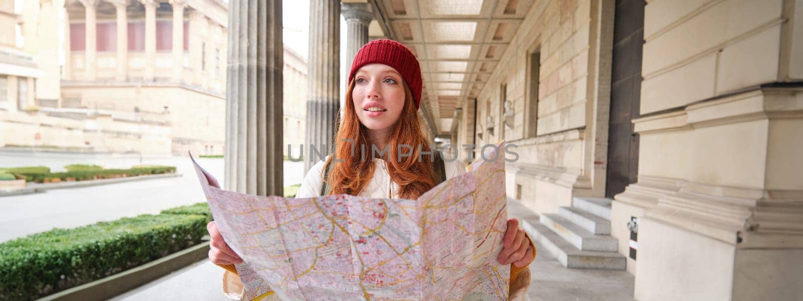 Adventurous redhead girl walks in town with paper map, explores city as tourist, looks for popular tourism attractions, looks around excited and smiles by Benzoix