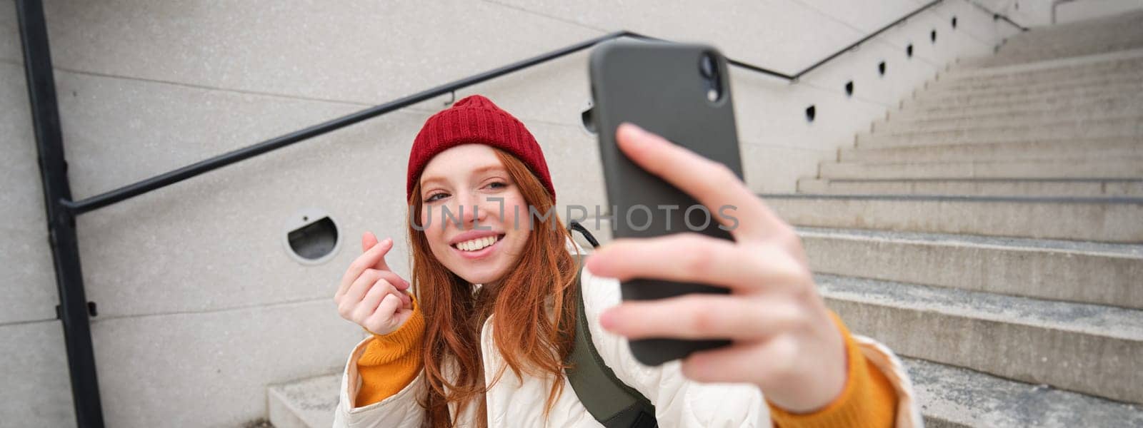 Mobile phone and people lifestyle. Stylish redhead girl takes selfie on her smartphone, poses for photo with mobile phone in hand, smiles happily, sits on stairs outdoors.
