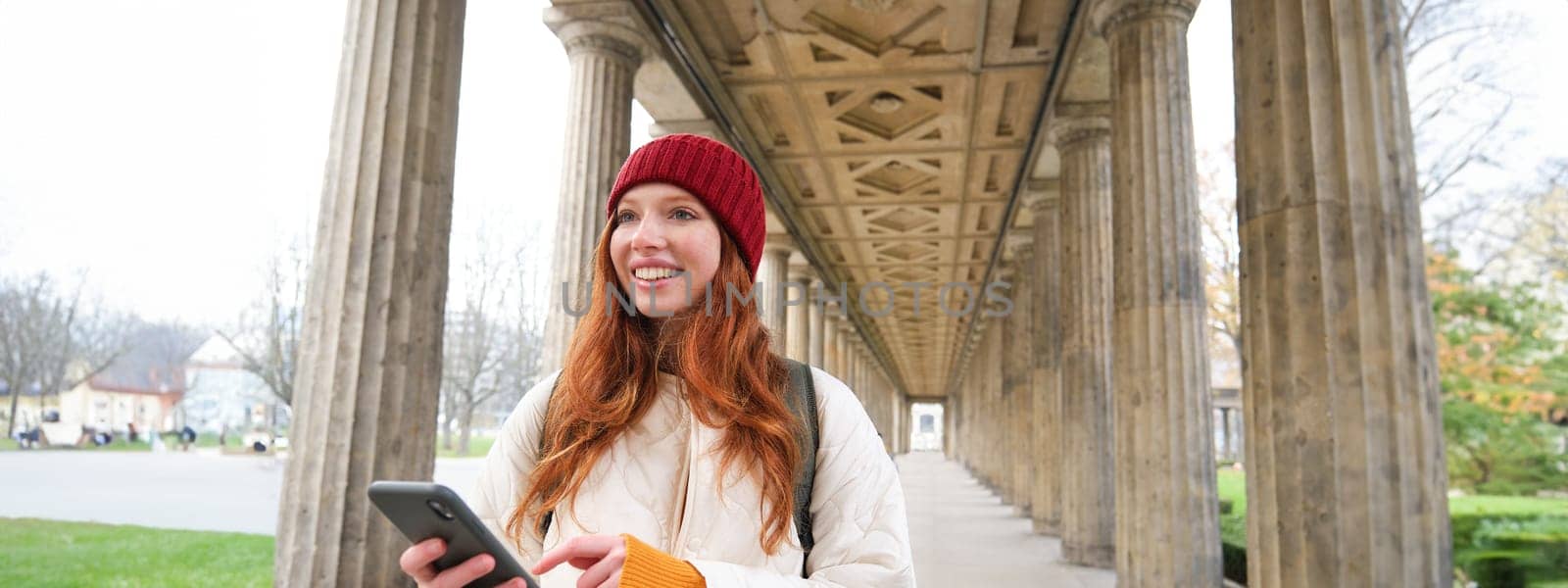 Happy young female model stands on street and holds mobile phone, uses smartphone app, follows the route of online map.