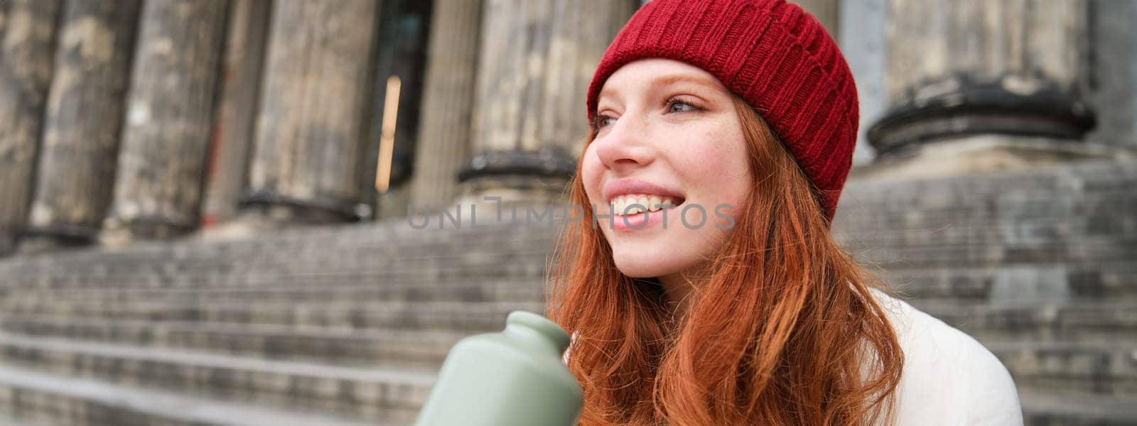 Smiling redhead girl in red hat, sits on stairs near building with flask, drinks water and looks happy by Benzoix