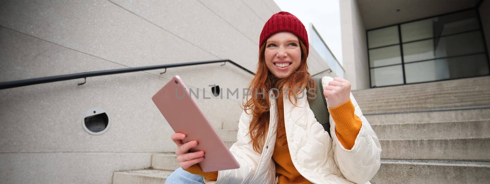 Happy stylish redhead girl, student in red hat, holds digital tablet, uses social media app, searches something online, connects to wifi by Benzoix
