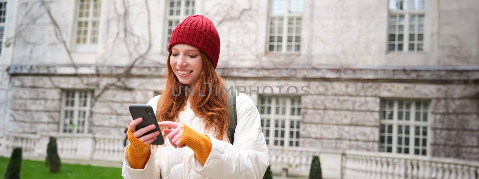 Portrait of young redhead woman looking at her mobile phone, checks her location on smartphone app, walking around city attractions.