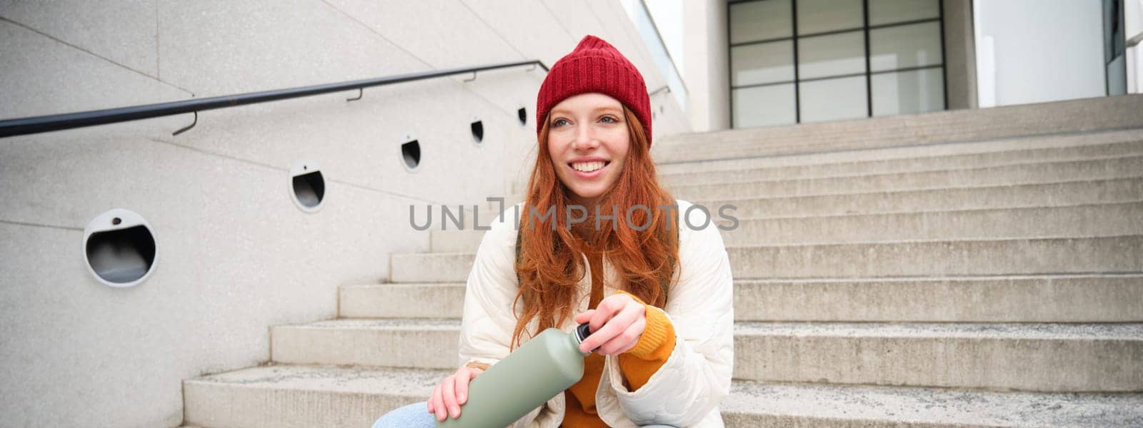 Smiling traveler, redhead girl tourist sits on stairs with flask, drinks hot coffee from thermos while travelling and sightseeing around foreign city, sits on stairs and rests.