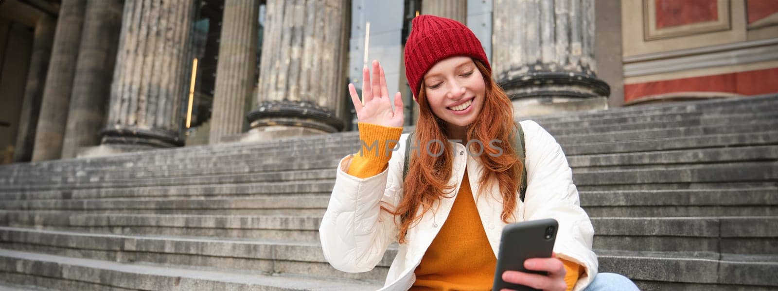 Redhead female tourist talks on mobile phone, video chatting with friends, says hi and waves hand at smartphone camera.