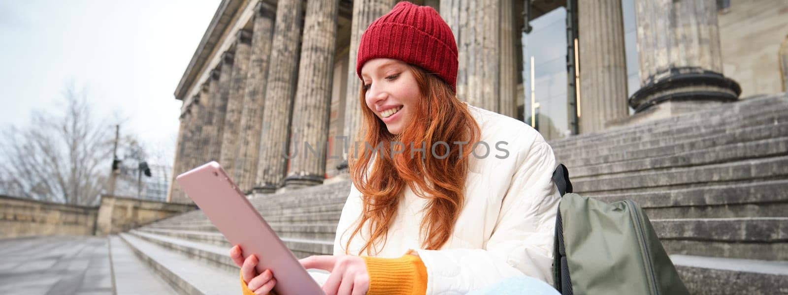 Beautiful young modern girl with red hair, holds digital tablet, sits on stairs near museum and connects public internet, sends message on gadget app.