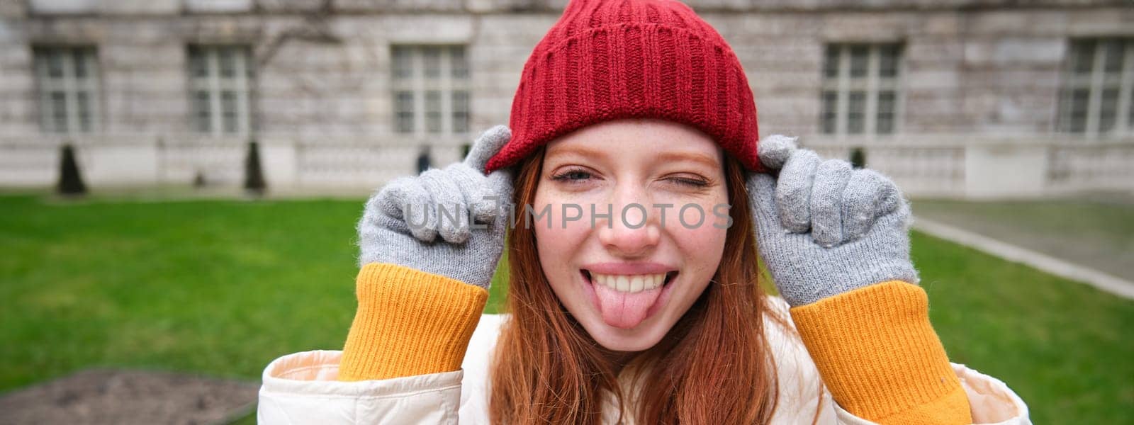 Portrait of funny and cute redhead girl puts on red hat, shows tongue and winks at camera, smiles happily, enjoys great weather in park.