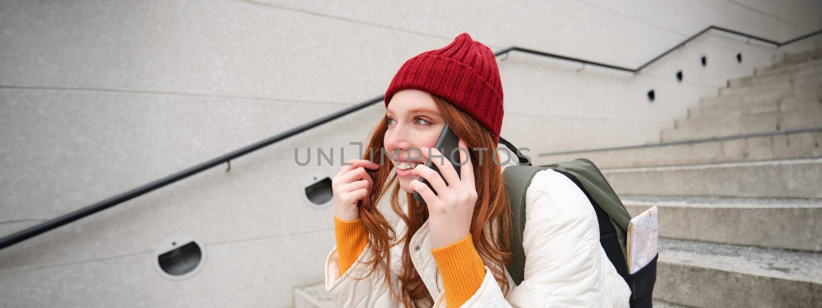 Beautiful smiling redhead female model, sits on street and talks on mobile phone, uses smartphone app to call abroad, laughing during telephone conversation by Benzoix