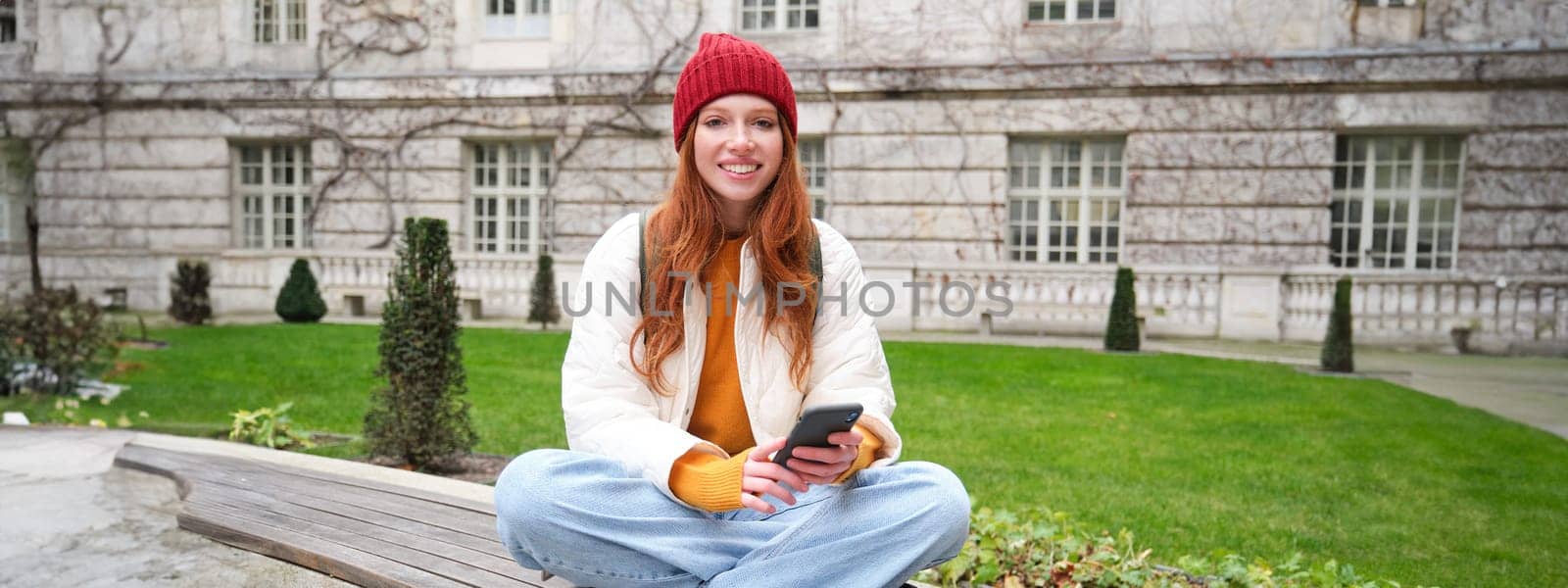 Portrait of stylish young woman, 25 years, sits on bench in park and uses mobile phone, reads online news, messages or watches video on smartphone app, connects to public wifi by Benzoix