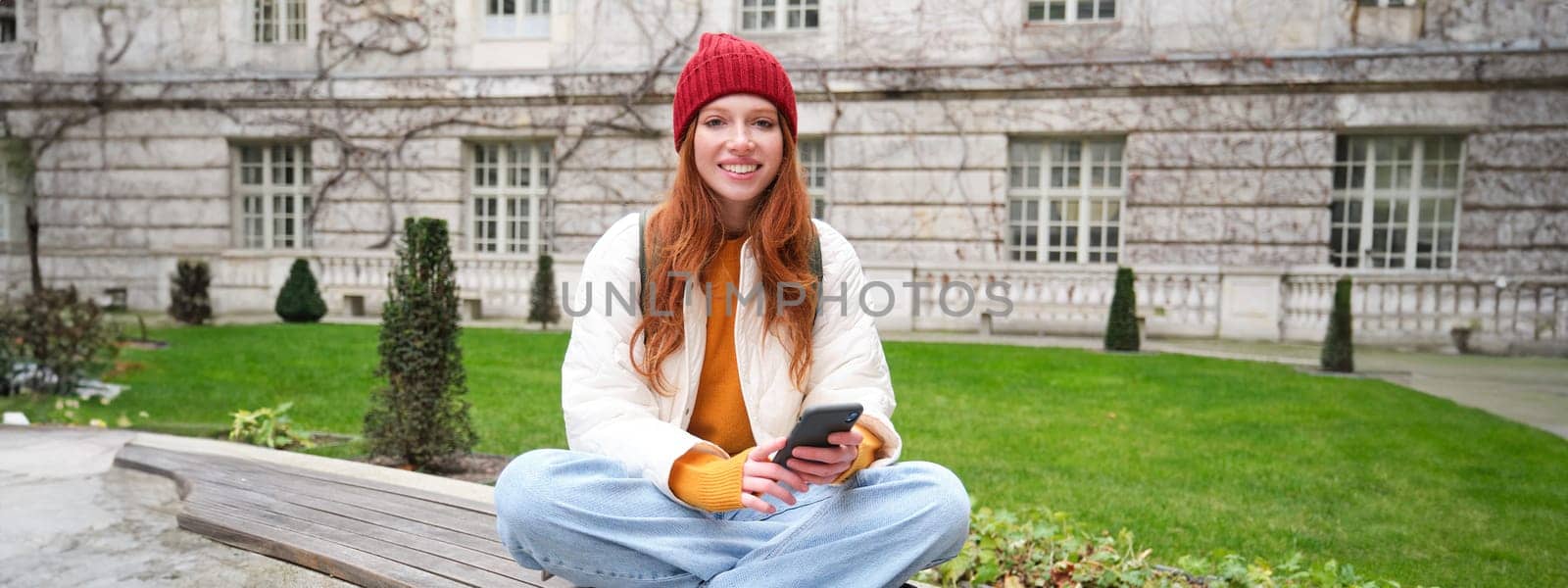 Portrait of stylish young woman, 25 years, sits on bench in park and uses mobile phone, reads online news, messages or watches video on smartphone app, connects to public wifi.