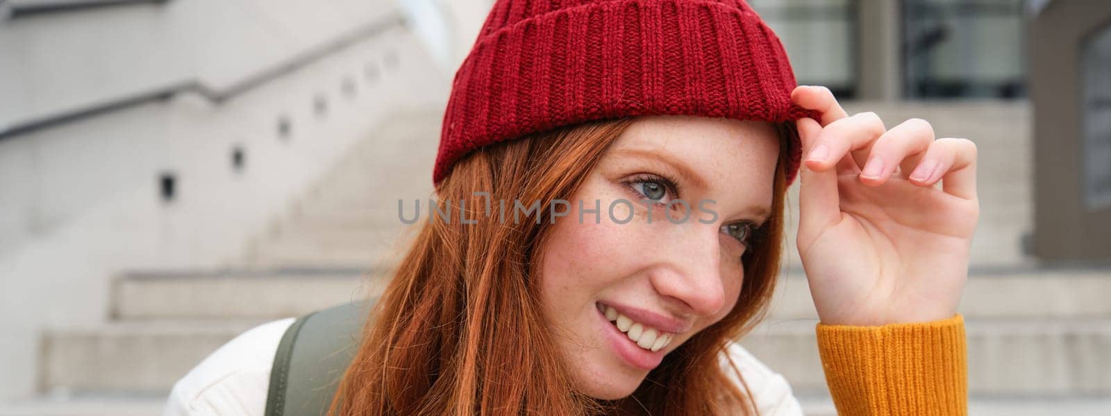 Stylish redhead girl in warm red hat, smiling relaxed, sitting with backpack on stairs near building, waits for someone outdoors by Benzoix
