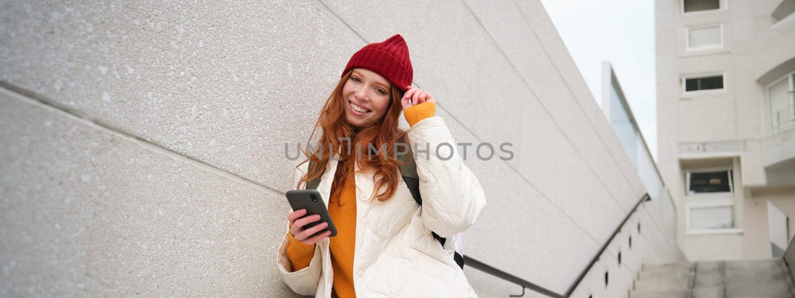 Happy girl student in red hat, holds smartphone, tourist looks at map app on her phone, explores sightseeing, texts message, looks for couchsurfing, rents place to stay online.