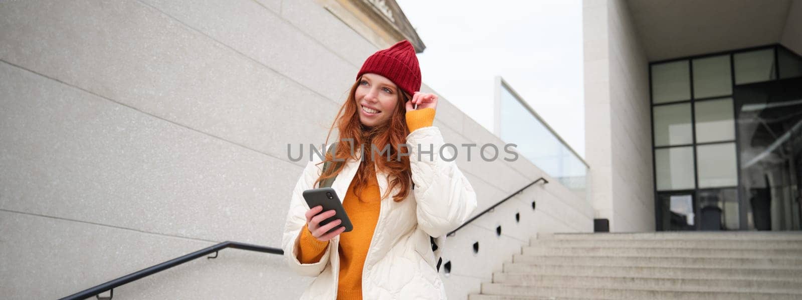 Happy girl student in red hat, holds smartphone, tourist looks at map app on her phone, explores sightseeing, texts message, looks for couchsurfing, rents place to stay online.