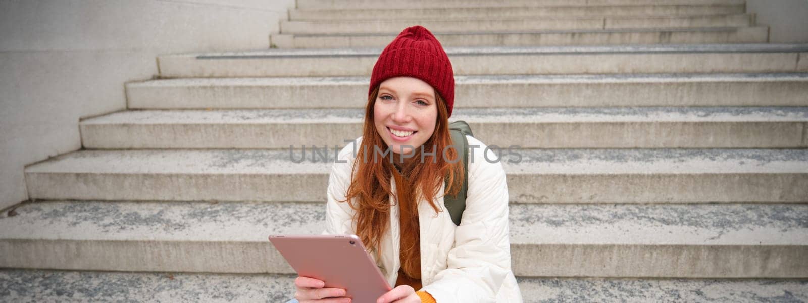 Happy stylish redhead girl, student in red hat, holds digital tablet, uses social media app, searches something online, connects to wifi by Benzoix