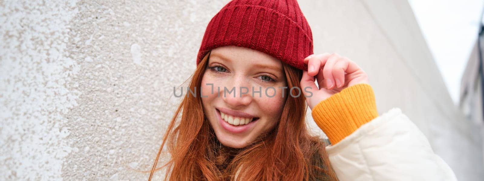 Stylish redhead girl in red hat, smiles and looks happy, poses outdoors on street, looks relaxed and lively.
