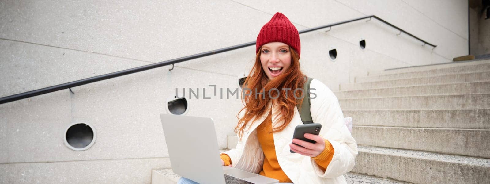 Young tourist, girl booking hotel room on laptop, enters confirmation code on smartphone, sitting with computer and phone on stairs outdoors by Benzoix