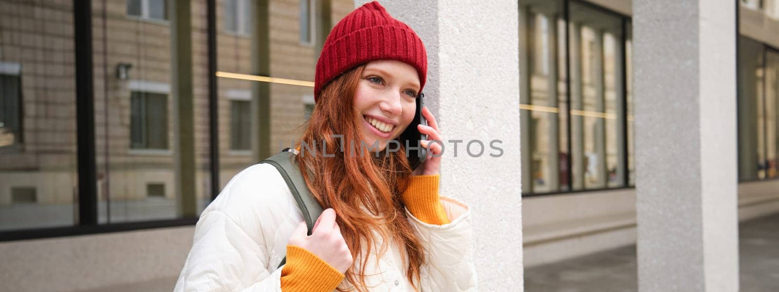 Portrait of smiling ginger girl with red hat, stands on street with backpack, rings someone on phone app, talks on mobile, uses smartphone.