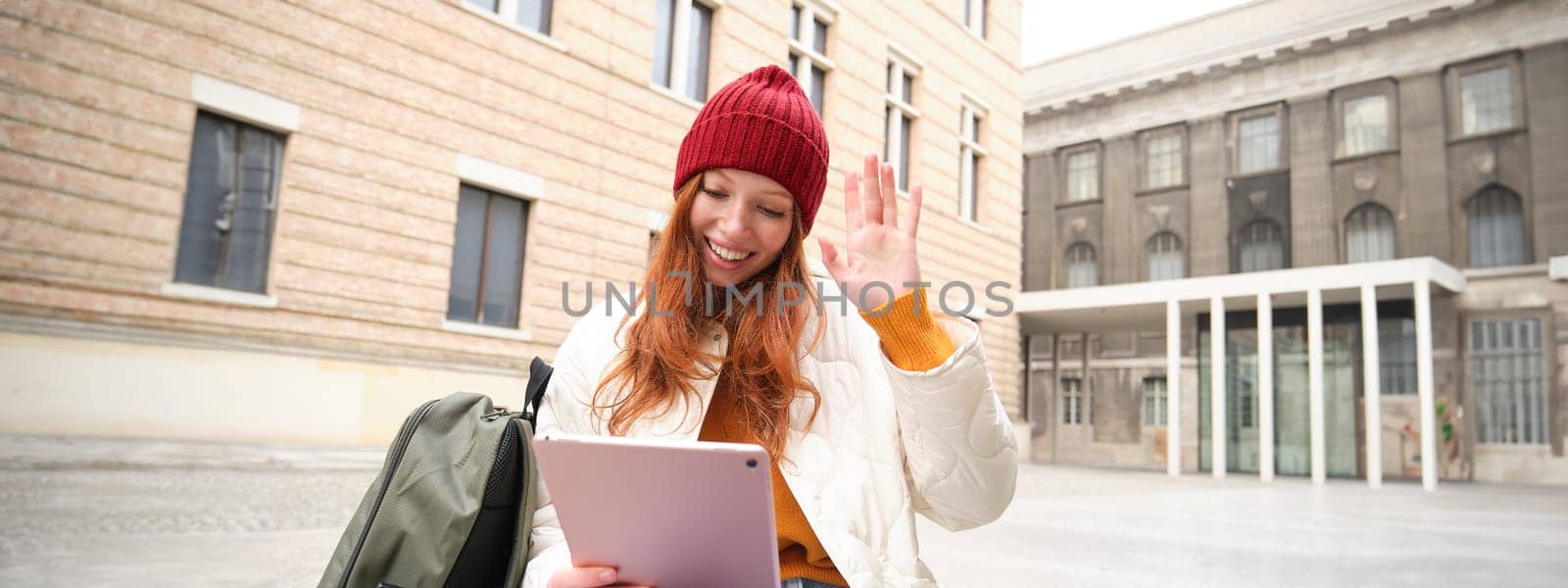 Happy redhead girl, student sits with backpack and digital tablet on street, says hello and waves at gadget camera, connects to public wifi to video chat, has online conversation.