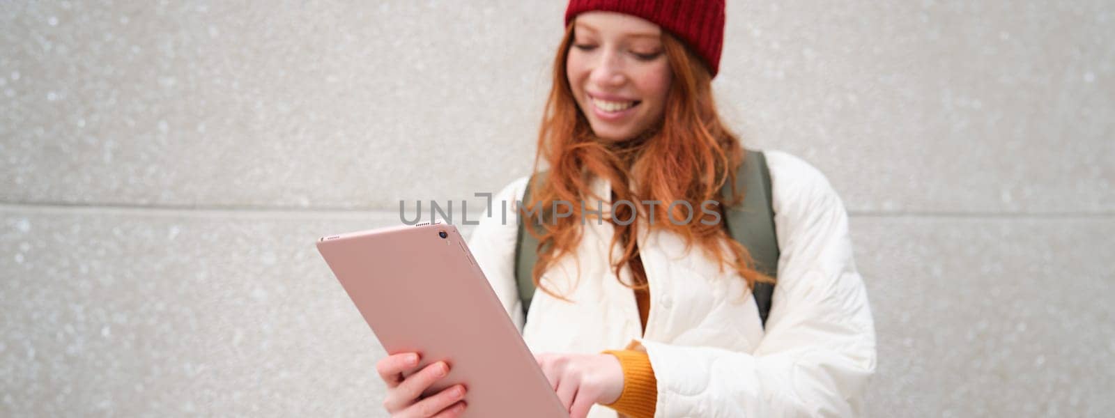 Young redhead girl, walks with digital tablet around city, stands on street with backpack and gadget, looks at online map, searches for a place in internet by Benzoix