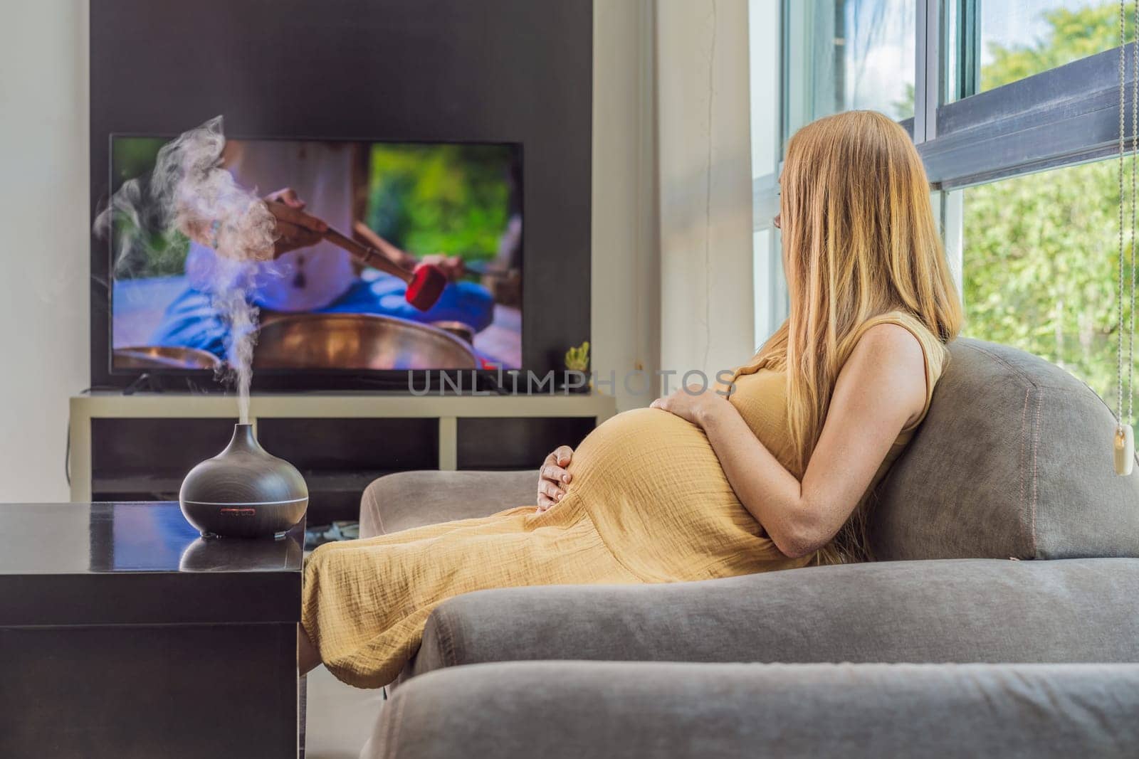 A blissful pregnant woman immerses in relaxation, savoring the soothing aroma from a diffuser while indulging in a calming TV video, embracing tranquility during her pregnant journey.