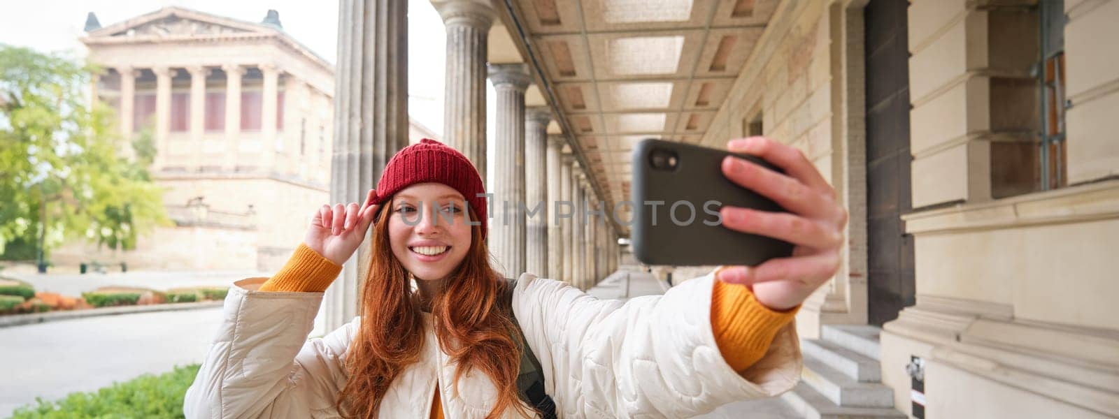Cute young redhead woman takes selfie on street with mobile phone, makes a photo of herself with smartphone app on street by Benzoix
