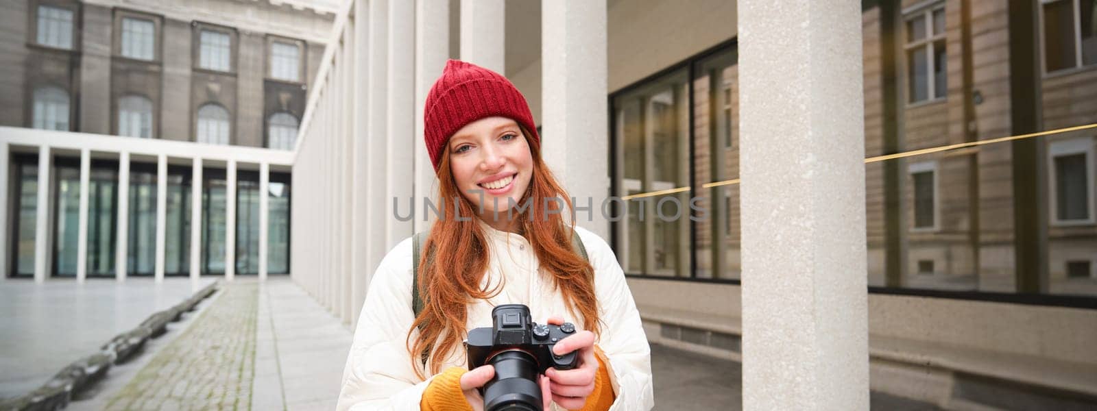 Smiling redhead girl photographer, taking pictures in city, makes photos outdoors on professional camera. Young talent and hobby concept