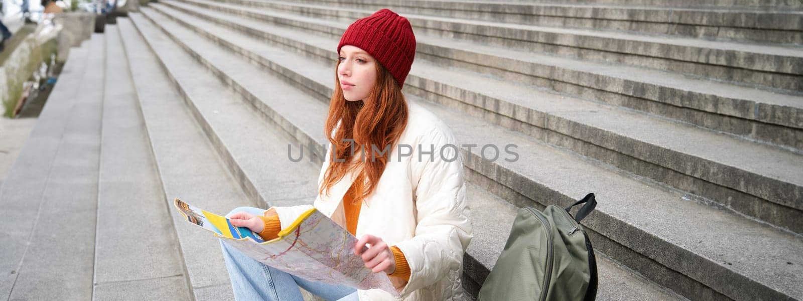 Tourism and lifestyle concept. Young redhead woman looking at city map, plans a route for sightseeing day, sits outdoors on stairs and rests.