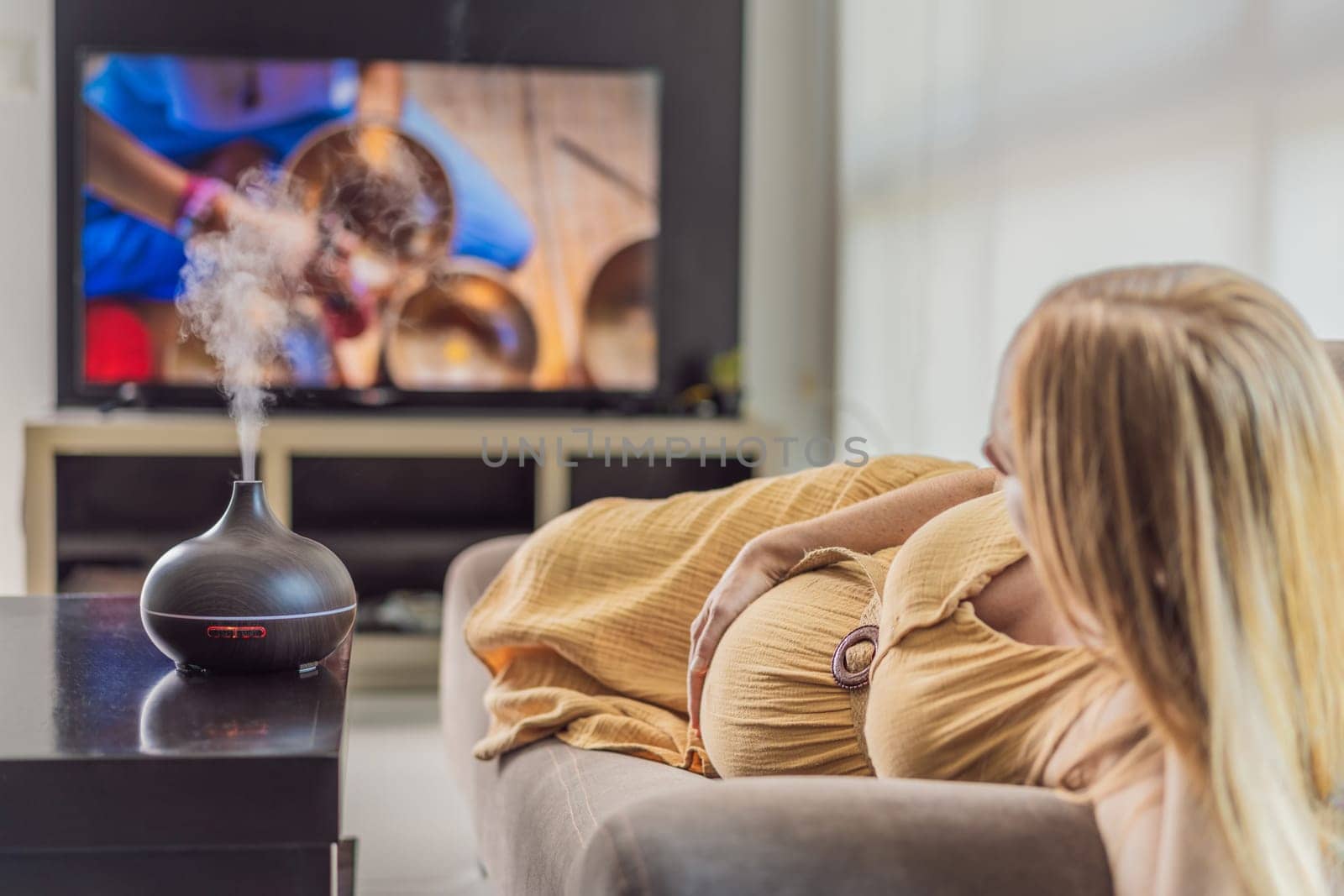 A blissful pregnant woman immerses in relaxation, savoring the soothing aroma from a diffuser while indulging in a calming TV video, embracing tranquility during her pregnant journey by galitskaya