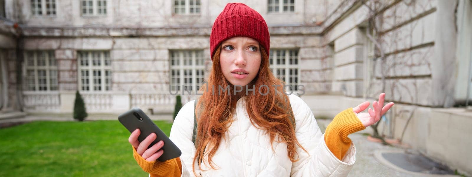 Portrait of confused girl tourist with smartphone, shrugs puzzled, dont know where to go, holds mobile phone with map application by Benzoix