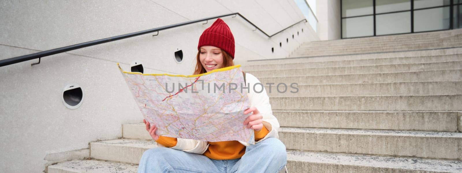 Beautiful girl tourist sits on stairs with city map, plans her journey, looks for direction while travelling around town, searches route for sightseeing.