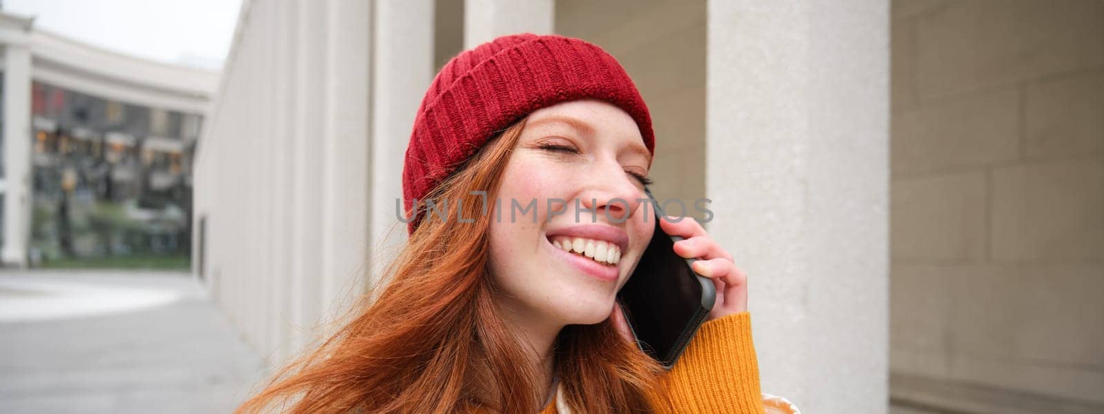 Mobile broadband and people. Smiling young redhead woman walks in town and talks on mobile phone, calling friend on smartphone, using internet to make a call abroad.