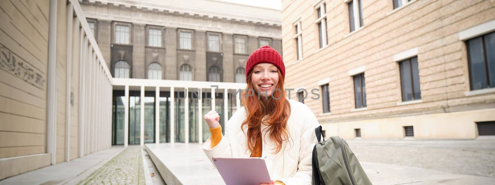 Beautiful redhead woman in red hat, sits with backpack and thermos, using digital tablet outdoors, connects to wifi, texts message, books tickets online.