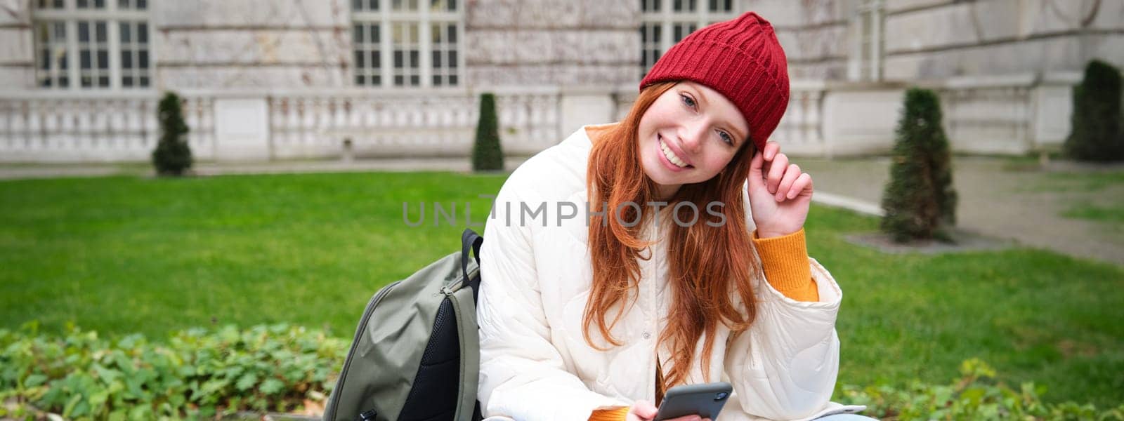 Young smiling redhead girl sits in park with smartphone, texting message on mobile phone, using telephone, connecting to public wifi and surfing internet.