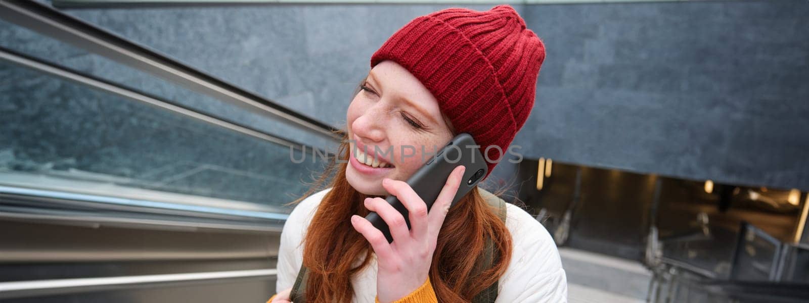 Portrait of happy redhead woman walking around town with smartphone, calling someone, talking on mobile phone outdoors.