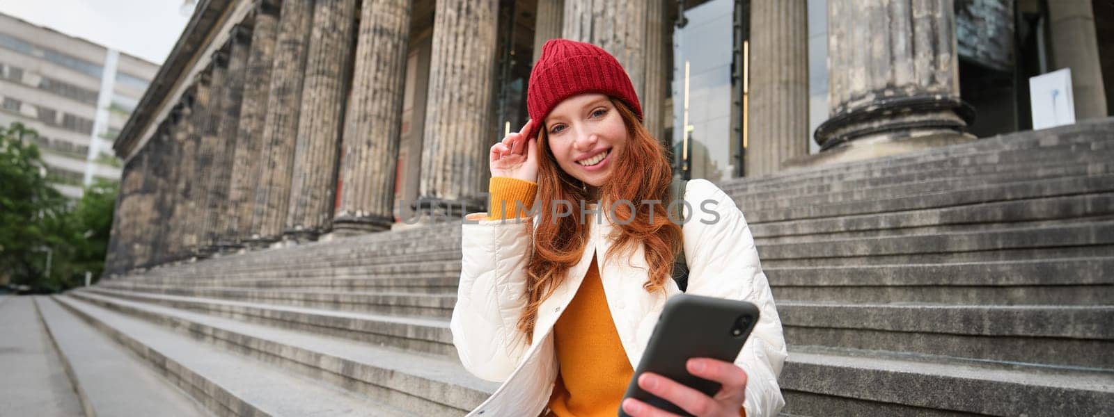 Portrait of young redhead woman in red hat, sitting on stairs, tourist looks at her mobile phone, rests on staircase of museum, connects public wifi.