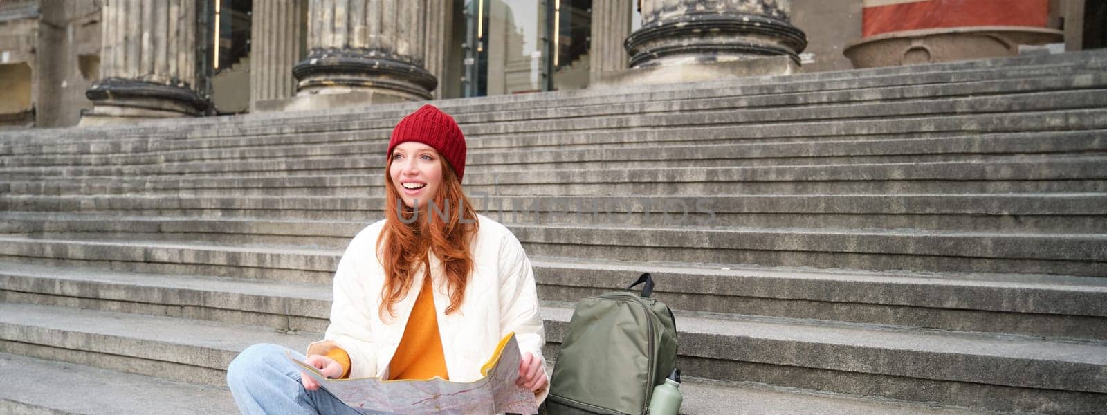 Portrait of young redhead woman, tourist sits with paper map and looks for a route to tourism attraction, rests on stairs outdoors.