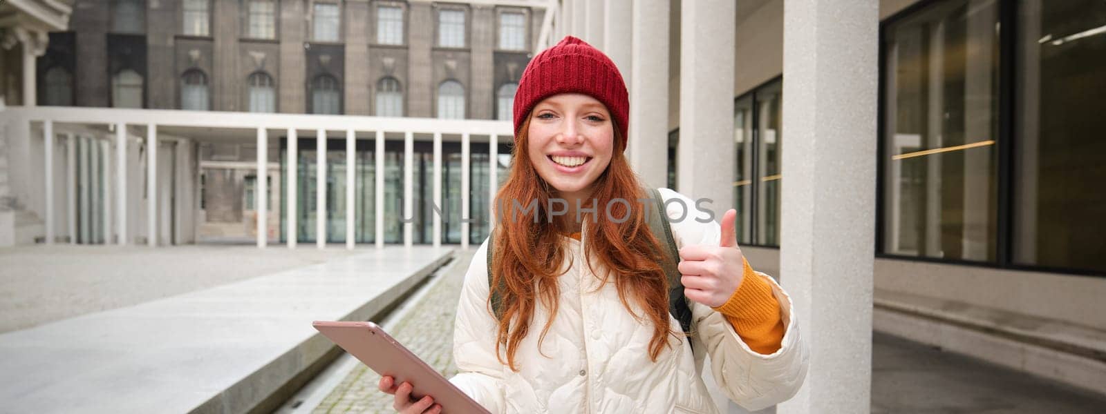 Happy redhead girl in red hat, walks around city with digital tablet, connects to public internet wifi and looks for route, looks at map on her gadget.